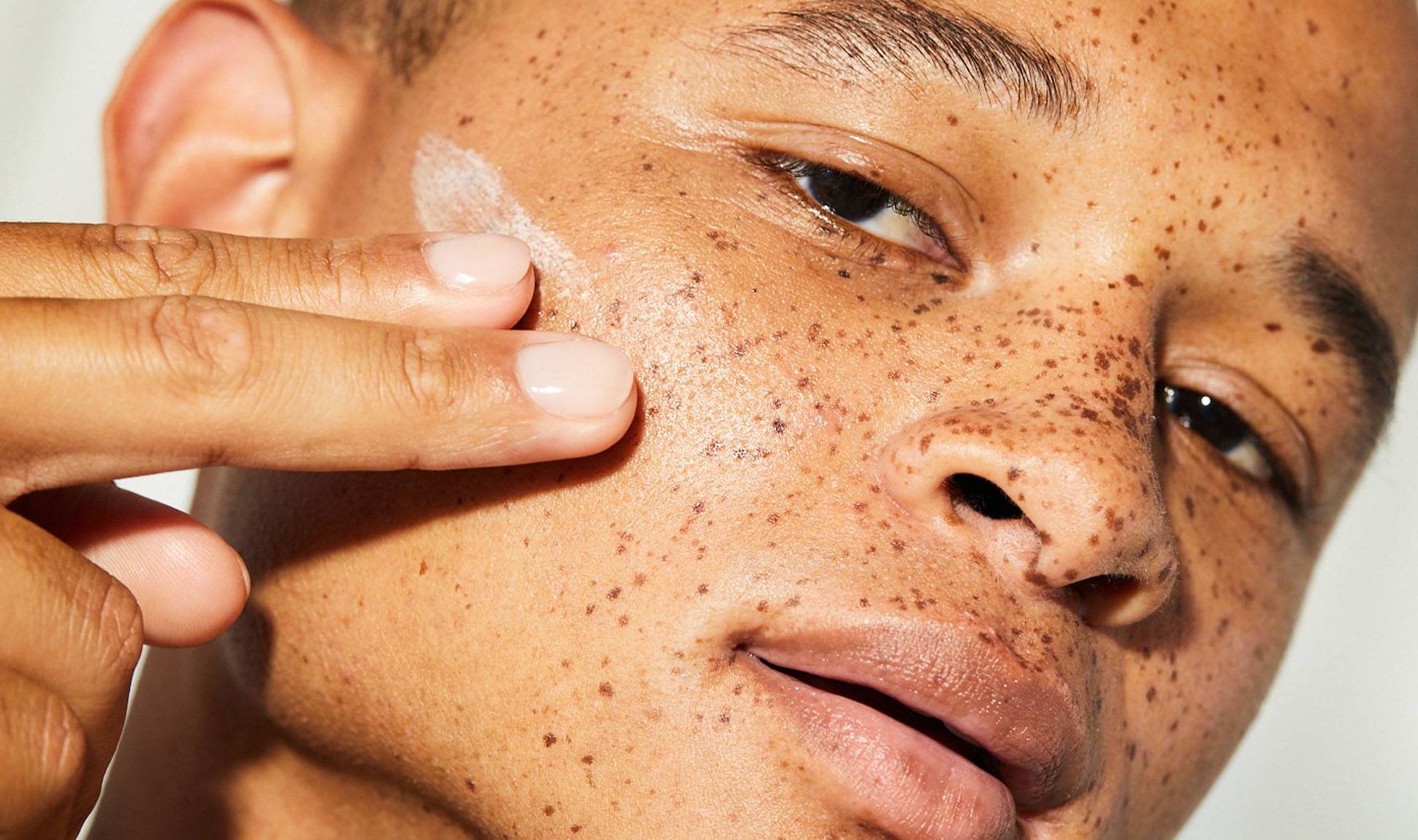 Closeup photo of a young man applying skincare product to his cheek with his fingers.