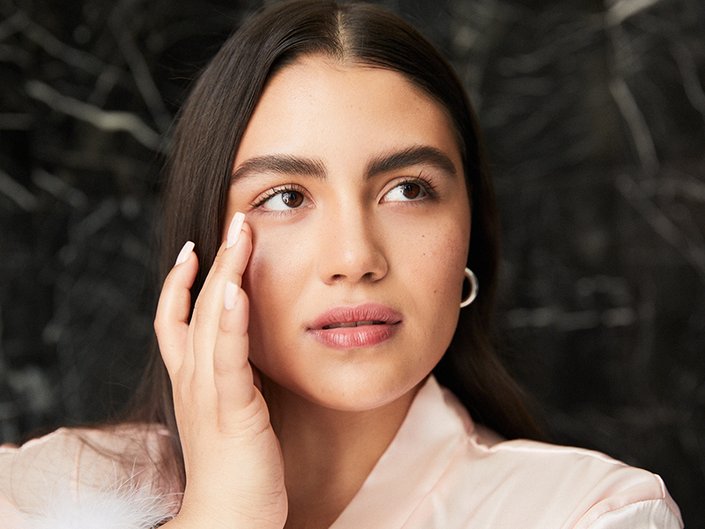 A person with long dark hair and white square-shaped nails touches their fingers to the outer corner of their eye and looks to the side of the camera
