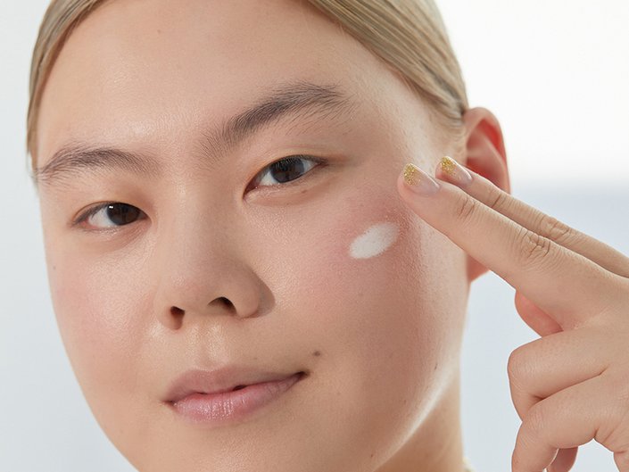 A person with their blonde hair pulled back looks at the camera and rubs face cream onto their cheek with two fingers
