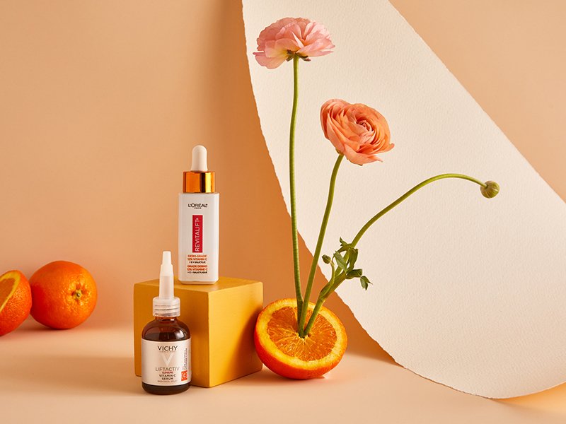 Image of two vitamin C serums on a neutral background next to oranges and orange flowers