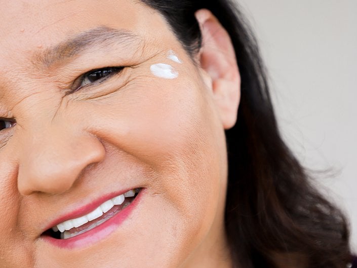 An older person with black hair smiling and a skincare product applied at the corner of their eye.