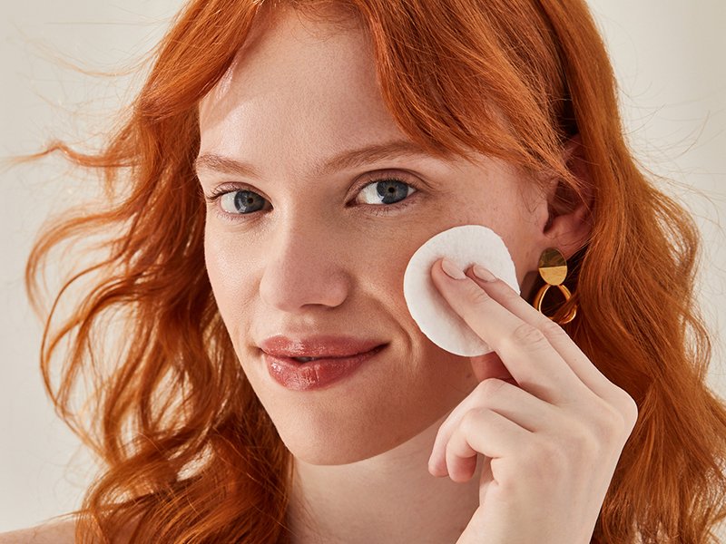 Person with red hair applying a cotton round to their face.