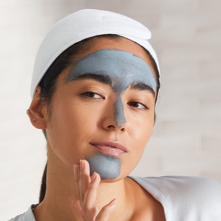 Image of a model applying a clay mask to her T-zone and chin, touching her chin