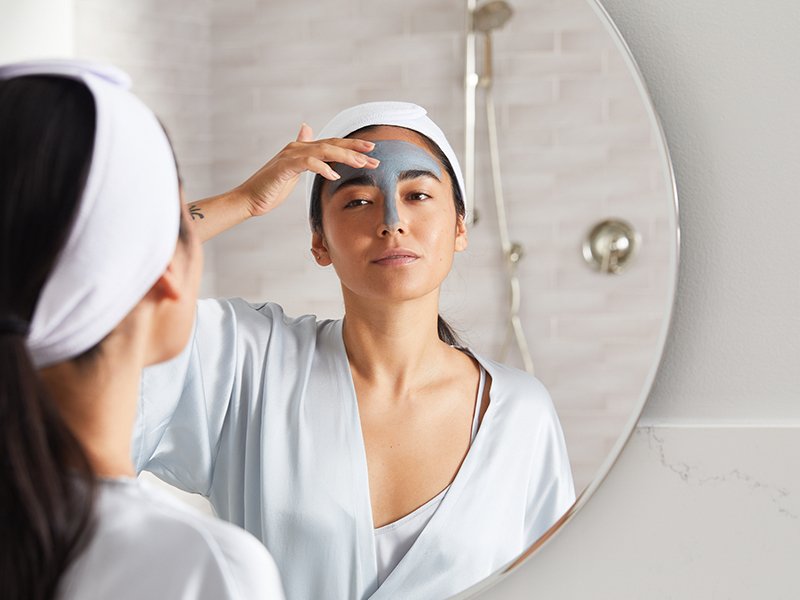 Image of model in a bathrobe, applying a clay mask to her T-zone while looking in the mirror