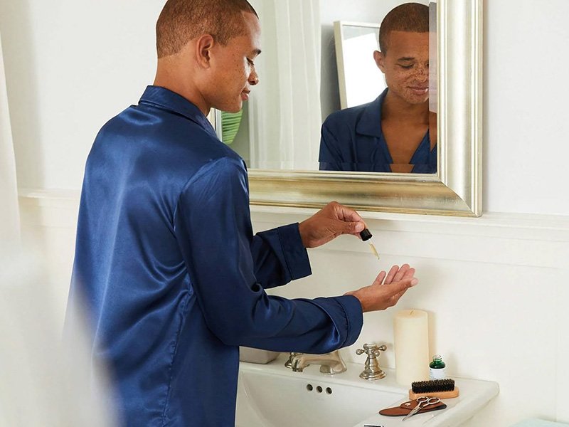 Person in blue pajamas standing at the bathroom sink applying a skincare product to their hand with a dropper