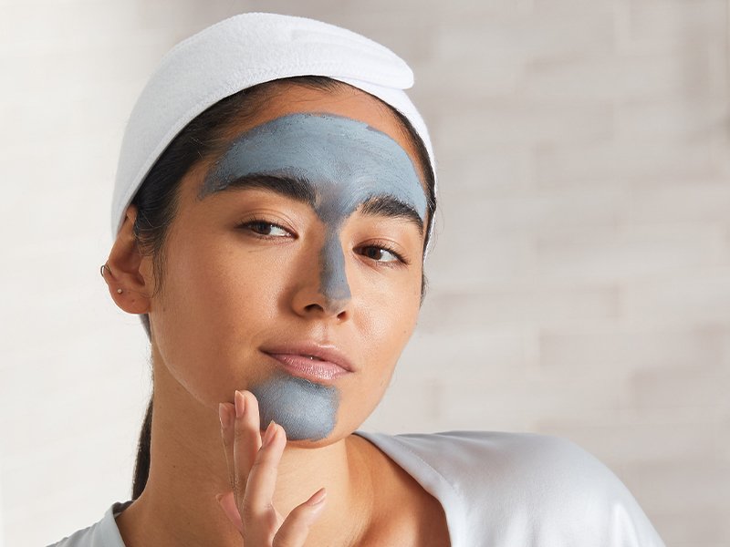 Person with a gray clay mask applied to their T-zone, staring in the mirror and touching their face.