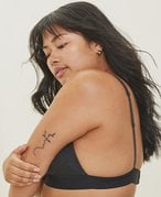 Person with long black hair wearing a black bra poses with their side facing the camera holding their arm. There is a tattoo on their arm.