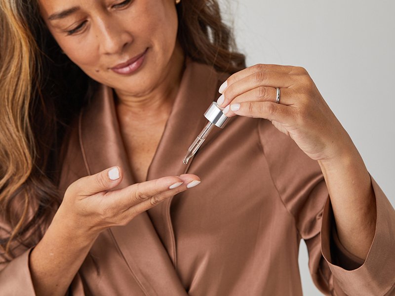 person in a brown silk robe using a dropper to apply serum to her fingertips 