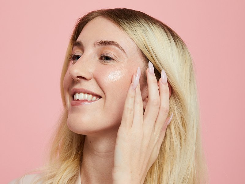 Photo of a blonde woman smiling and applying moisturizer to her cheek