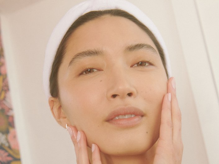 Close-up picture of a model looking at the camera and touching the sides of their face while wearing a towel wrapped around their head