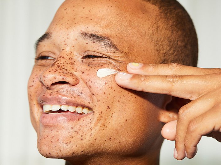 Person with freckles smiling and swatching face cream on their cheek with two fingers
