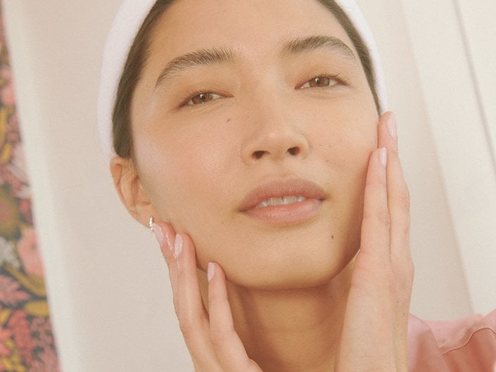 Picture of a model smiling softly while touching the sides of their face and looking at the camera with a towel wrapped around their head