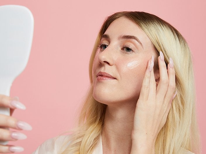 Person with blonde hair touching her fingers to her cheeks and looking in a hand-held mirror