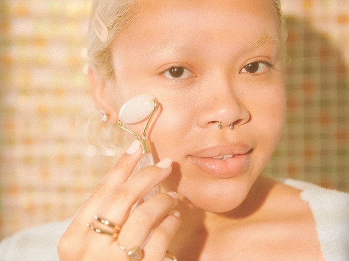 Person with blonde hair using a face massager on their cheek and looking at the camera