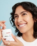 Person with dark curly hair smiling and holding a Thayers moisturizer