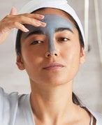 Model with brunette hair pulled back into a ponytail, applying a gray charcoal face mask to their T-zone.