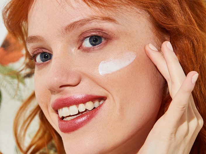 Picture of a model with red hair smiling at the camera and applying a white smear of sunscreen to their face.