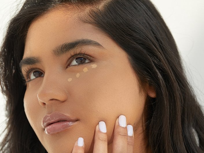 Close-up of person's face with foundation swatched in dots on their cheek. Person has long dark hair and is holding her finger to her jaw.
