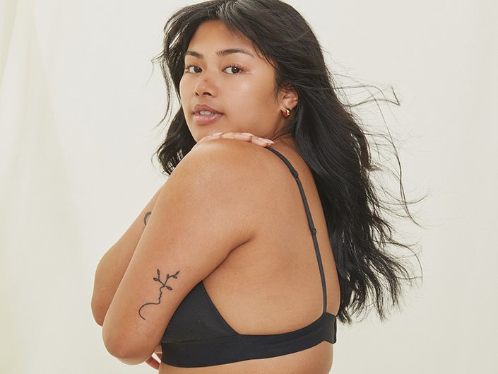 Person with long dark hair and clean skin with no makeup in a black bra top is standing facing the side, hugging their arms around themself and looking at the camera.