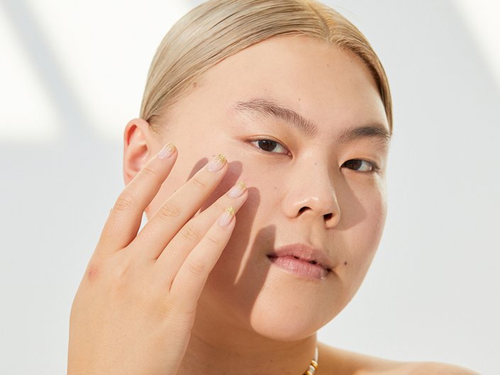 Person with blonde hair pulled back looking at the camera and touching their fingertips to their cheek