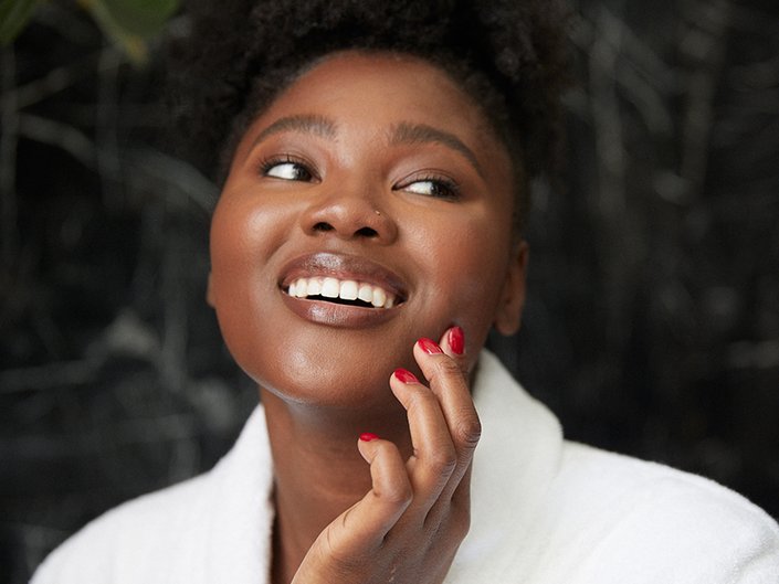 A person smiling and looking away from the camera touches their hand with red painted nails to their cheek