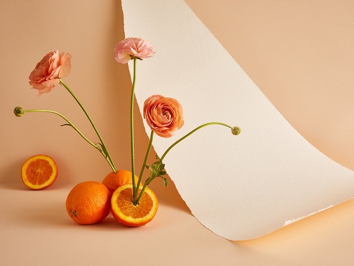 Whole oranges and oranges sliced in half with orange flowers growing out of them, against a peach-colored background