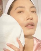 Person wearing a white headband and pink satin robe blotting their cheek with a white washcloth 