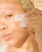 Close-up picture of a model with bleached eyebrows and a septum piercing, holding a white gua sha to their cheek in the sunlight