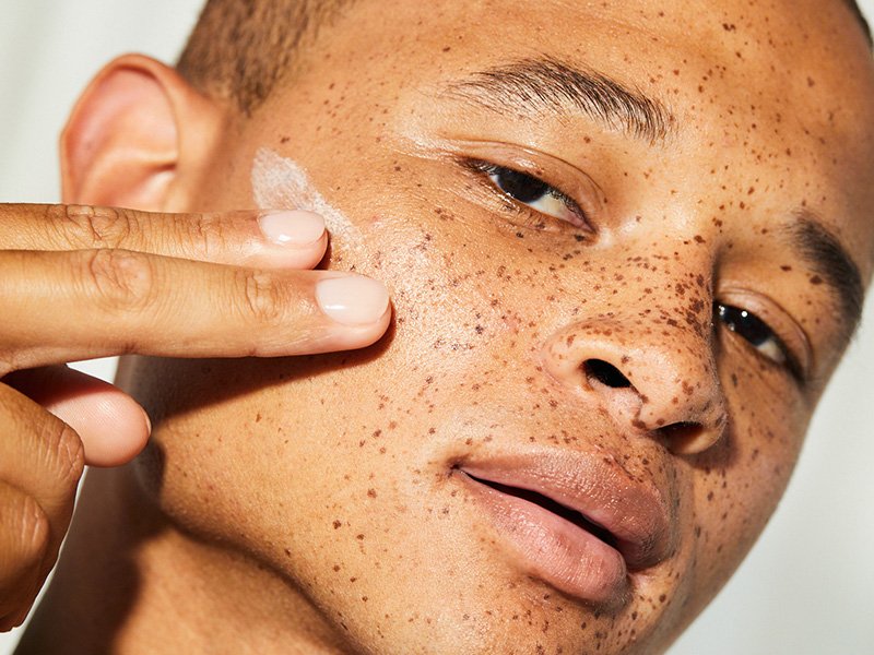 Close-up picture of a male model with freckles applying sunscreen to his cheek