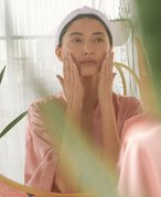 person in a pink satin robe and white headband looks in the mirror and touches their hands to their cheeks