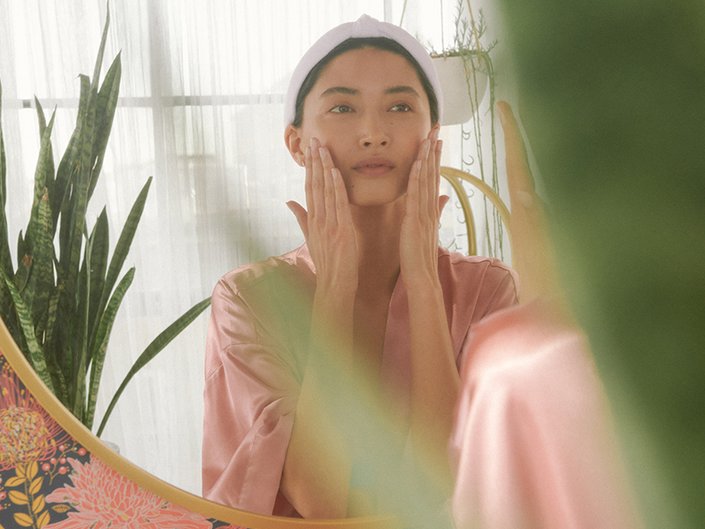 person in a pink satin robe and white headband looks in the mirror and touches their hands to their cheeks