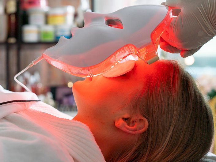 A person lying on their back in a white spa robe having an LED light mask placed on their face
