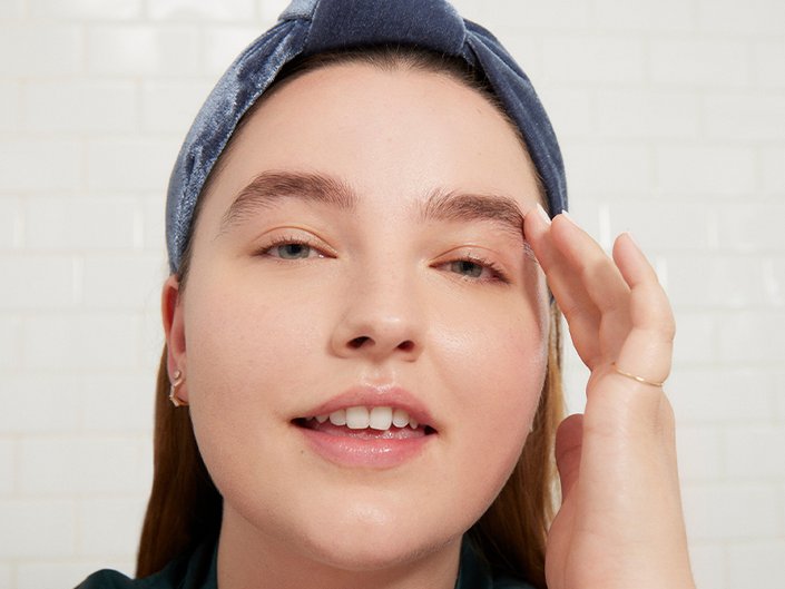 Close-up picture of a brunette model softly smiling and touching the side of their face, wearing a blue headband
