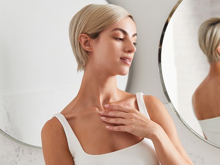 person with a blonde pixie cut wearing a white tank top and holding their hand to their neck