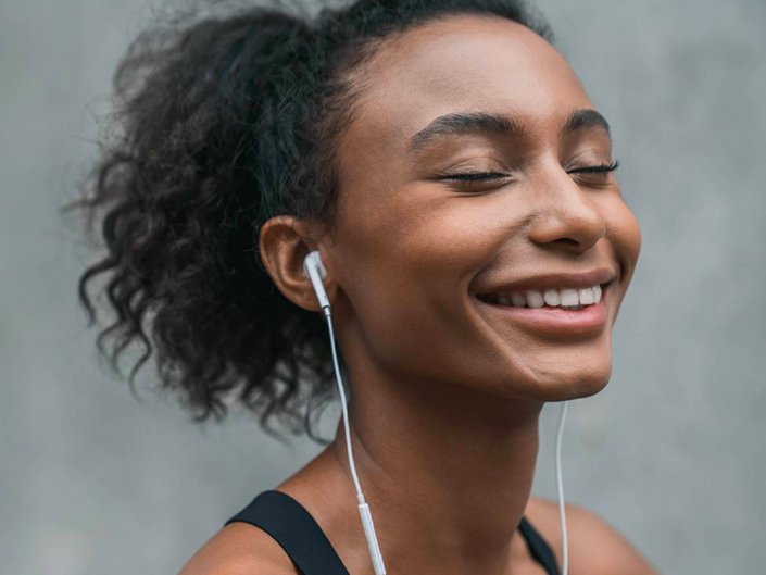 Person with dark hair pulled back in a ponytail smiling with their eyes closed and wearing earphones