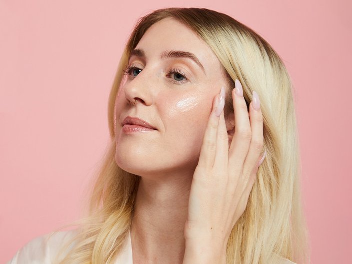 Picture of a blonde person putting moisturizer on their cheek, on a light pink background