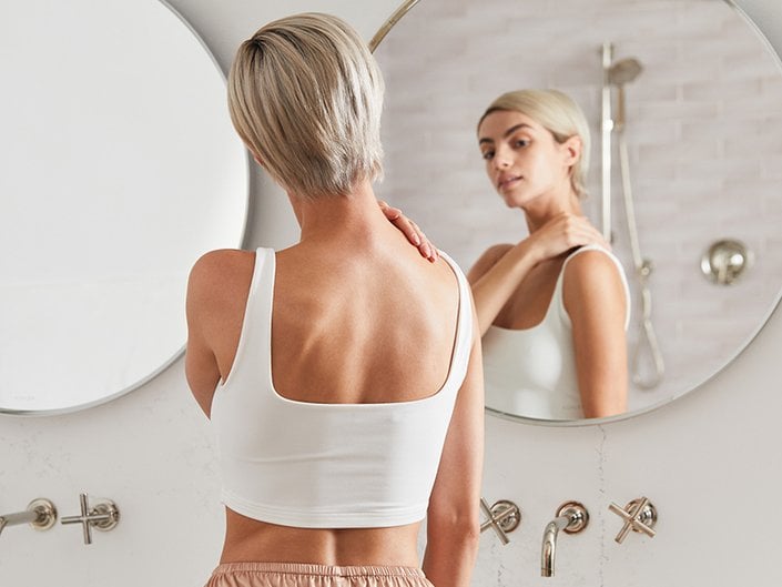 Person with a blonde pixie cut wearing a white tank top and looking in the bathroom mirror while touching their shoulder