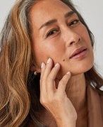 Person with long dark hair and brown eyes touching their fingers to their cheek