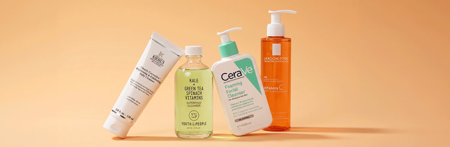 Bottles of Kiehl’s Clearly Corrective Brightening & Exfoliating Daily Cleanser, Youth to the People Kale + Green Tea Spinach Vitamins Superfood Cleanser, CeraVe Foaming Facial Cleanser, and La Roche-Posay Vitamin C Brightening Cleanser on an orange background