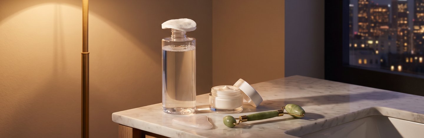 A bottle of clear micellar water with a cotton pad, on a bathroom vanity beside a moisturizer and skincare tools, in soft evening lighting